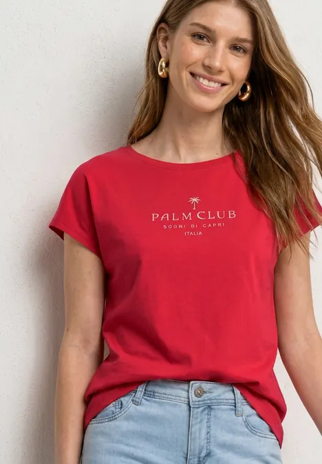 Vrouw draagt een rood T-shirt met Palm Club Côte d'Azur opdruk en een lichtblauwe jeans voor een witte muur.