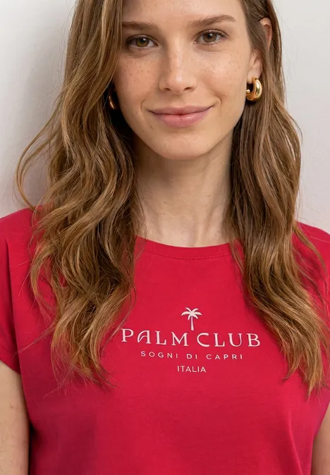 Persoon met rood T-shirt «PALM CLUB FOOD & GRACE ITALIA» en halflang golvend haar tegen een witte achtergrond