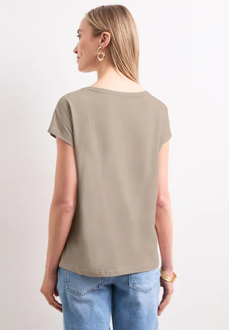 Shirt met hangende schouder en tekst clay sand Vrouw staat met de rug naar de camera, draagt een kaki t-shirt en een blauwe spijkerbroek.