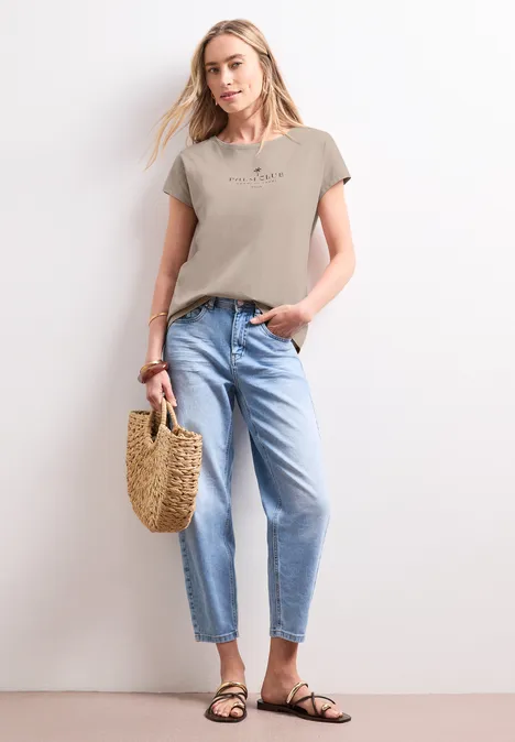 Shirt met hangende schouder en tekst clay sand Vrouw in beige T-shirt, lichtblauwe jeans, sandalen en rieten tas.