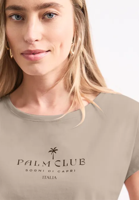 Shirt met hangende schouder en tekst clay sand Persoon draagt beige t-shirt met opdruk 'Palm Club Sogni di Capri Italia' en palmbomenmotief.