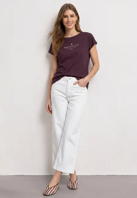 Een vrouw staat voor een muur in witte jeans, paars T-shirt en gestreepte pumps.