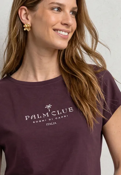 Vrouw met golvend haar, gouden bloemenoorbellen en donkerpaars t-shirt met 'Palm Club'-opdruk'