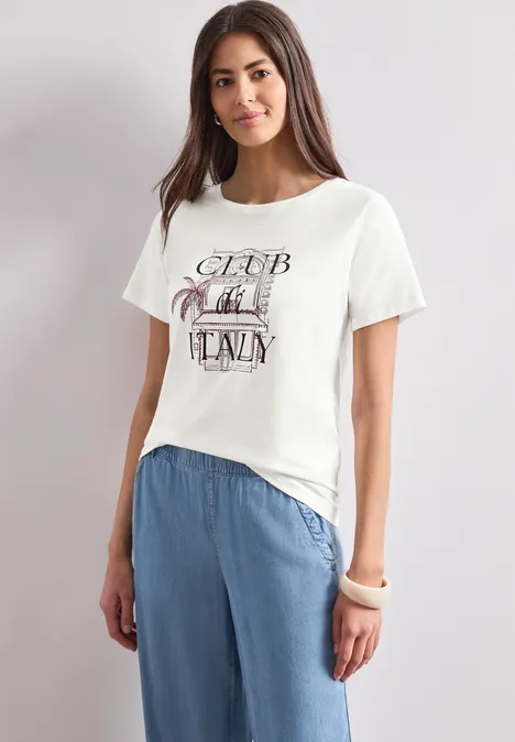 T-Shirt mit Rundhals und Artwork off white Eine Frau trägt ein weißes T-Shirt mit Aufdruck und lockere, hellblaue Hose vor neutralem Hintergrund.