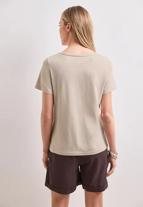 Shirt met ronde hals en Artwork clay sand Achteraanzicht van een blondine in beige T-shirt en bruine korte broek voor een lichte achtergrond.