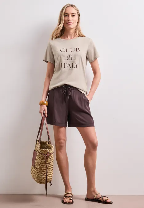Shirt met ronde hals en Artwork clay sand Vrouw in zomerse casual kleding: t-shirt, korte broek, sandalen, met een strooien strandtas.