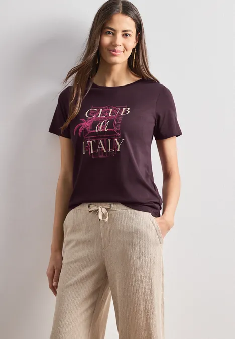 Frau in lockerem Outfit: dunkles T-Shirt mit 'Club Italy'-Schriftzug und helle Leinenhose.