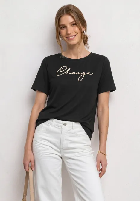 Shirt met ronde hals en Wording Artwork Black Vrouw met zwart T-shirt 'Change' en witte broek, draagt een tas, voor een lichte achtergrond.