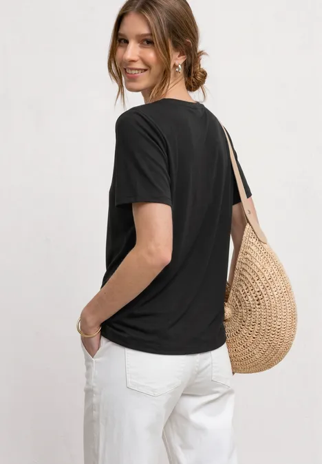 Shirt met ronde hals en Wording Artwork Black Vrouw van achteren in zwart t-shirt en witte broek met een gevlochten strooien tas.