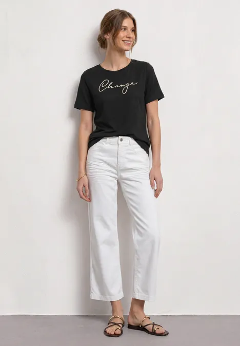 Shirt met ronde hals en Wording Artwork Black Een vrouw in een zwart T-shirt, witte broek en platte sandalen voor een lichte achtergrond.