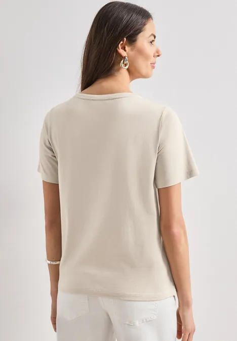 T-shirt à col rond avec wording artwork clay sand Femme de dos en t-shirt beige et pantalon blanc, cheveux bruns mi-longs.