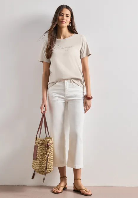 T-shirt à col rond avec wording artwork clay sand Femme en haut beige, pantalon blanc large et sandales, portant un sac en paille sur fond clair