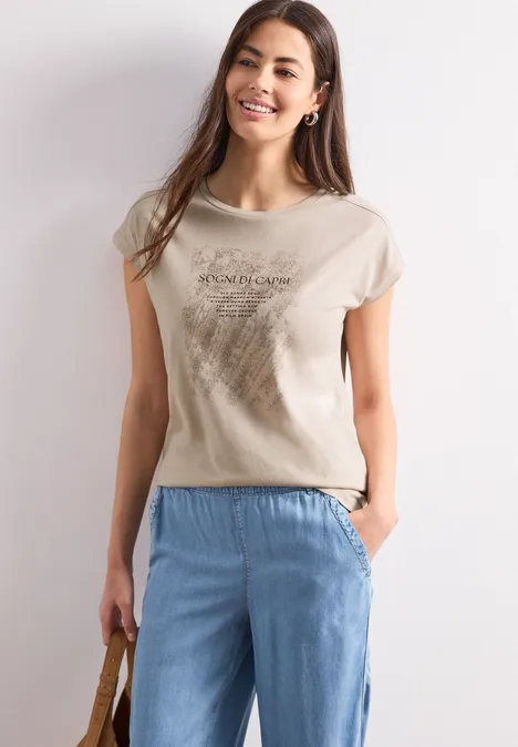 Chemise dropped shoulder avec artwork clay sand Une femme porte un t-shirt beige et un pantalon bleu clair, tenant un sac à la main.