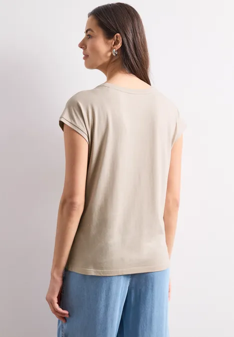 Chemise dropped shoulder avec artwork clay sand Femme vue de dos portant un T-shirt beige à manches courtes et un jean ample bleu, fond neutre.