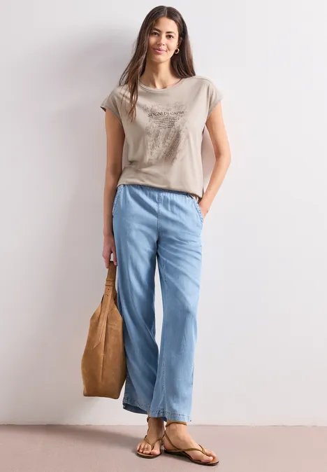 Chemise dropped shoulder avec artwork clay sand Femme décontractée portant un t-shirt beige, un jean ample bleu, des sandales et un sac à main.