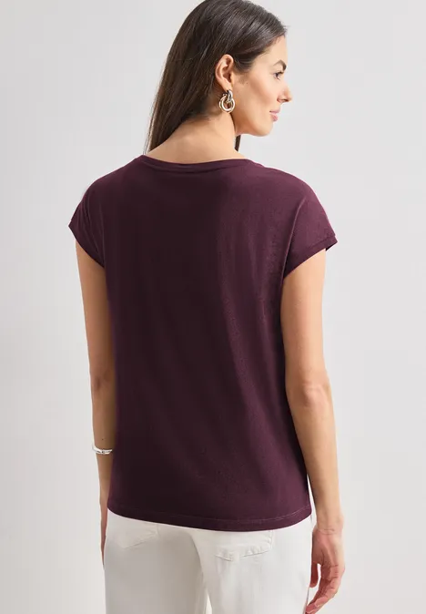 Frau von hinten in violettem T-Shirt und weißer Hose, lange Haare, Ohrringe und Armband.