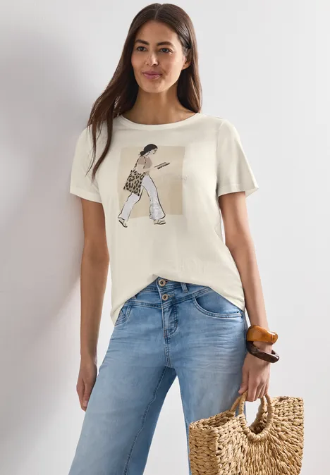 Frau in beigem T-Shirt mit Print, blauen Jeans und geflochtenem Strohtäschchen in lässiger Pose.