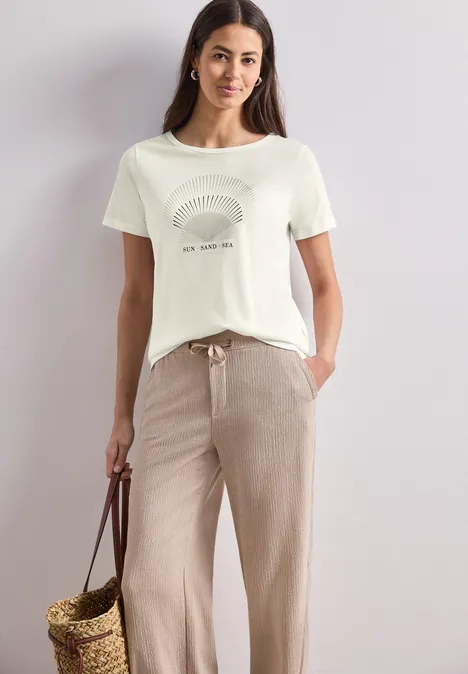 T-Shirt mit Rundhals und Foliendetails off white Frau in weißem T-Shirt mit Muster, beiger Hose und geflochtener Korbhandtasche.