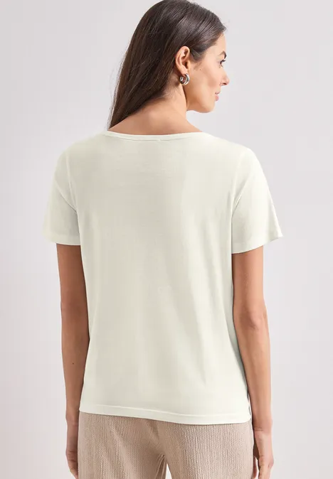 T-Shirt mit Rundhals und Foliendetails off white Rückenansicht einer Frau in schlichter cremefarbener Kurzarmbluse und beiger Hose vor hellem Hintergrund.