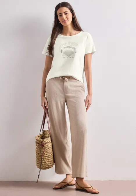 T-Shirt mit Rundhals und Foliendetails off white Frau in beigen Leinenhosen, weißem Print-T-Shirt, Strohtasche und Sandalen vor neutralem Hintergrund.