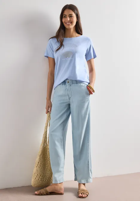 Shirt met ronde hals en foliedetails cashmere blue Vrouw in lichtblauwe wijde jeans, pastelblauw t-shirt en grote strooien tas voor een witte muur.