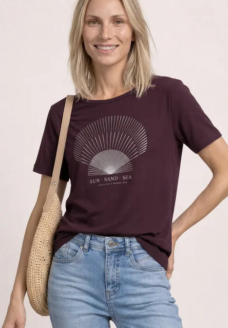 Vrouw in paars shirt met schelpenprint en jeans, draagt een gevlochten tas.