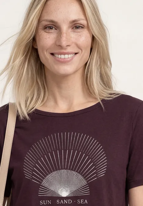 Vrouw met lichtblond haar draagt een donkerpaars shirt met zonmotief en de tekst 'Sun Sand Sea'.