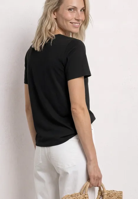 T-Shirt mit Rundhals und Embroidery Detail Black Person von hinten in schwarzem T-Shirt und weißer Hose, hält eine Strohtasche.