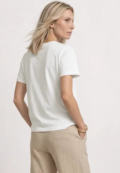 Shirt met ronde hals en geborduurd detail off white Achteraanzicht van een vrouw met een los wit T-shirt en beige broek voor een lichte achtergrond.