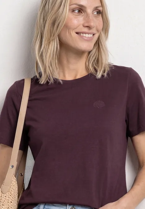 Eine lächelnde Frau in kastanienbraunem T-Shirt mit beiger Tasche vor hellem Hintergrund.