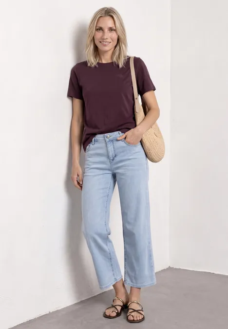 Frau lehnt an weißer Wand, trägt lila T-Shirt, helle Jeans, schwarze Sandalen und Korbtasche.