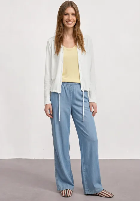 Lichtgewicht Langam-jack in linnenlook off white Een vrouw draagt een witte jas, geel shirt, losse lichtblauwe broek en gestreepte sandalen tegen effen achtergrond.