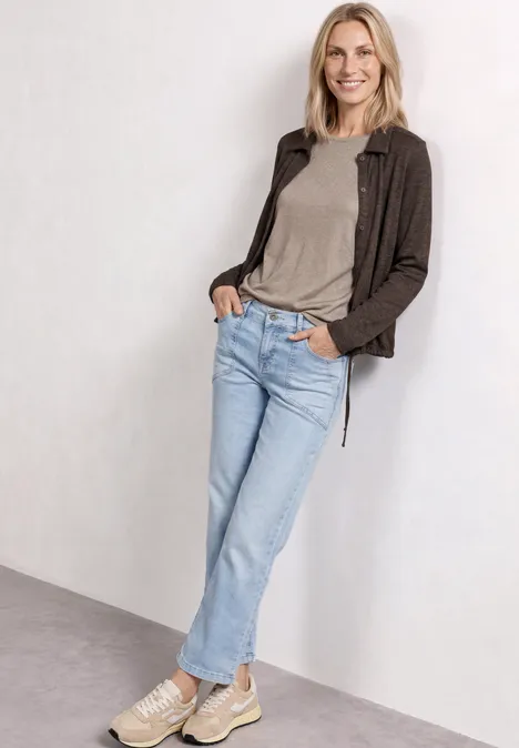 Femme posant en tenue décontractée: t-shirt gris, veste marron, jean bleu clair et baskets blanches.