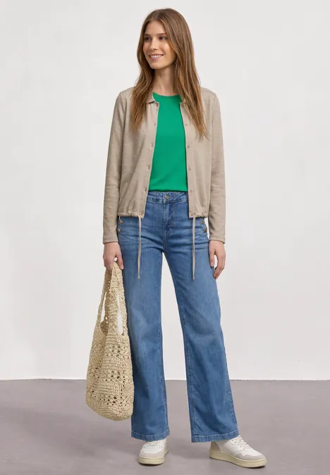 Junge Frau trägt grünes T-Shirt, helle Jacke, blaue Jeans, weiße Sneaker und eine gehäkelte Tasche.