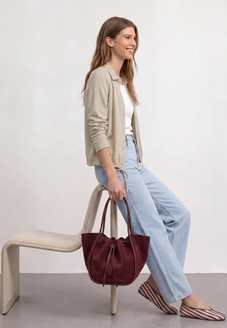 Leichte Langam Jacke in Leinen-Optik timeless beige Eine Frau sitzt seitlich auf einer Bank, trägt Cardigan, Jeans und hält eine dunkelrote Handtasche.