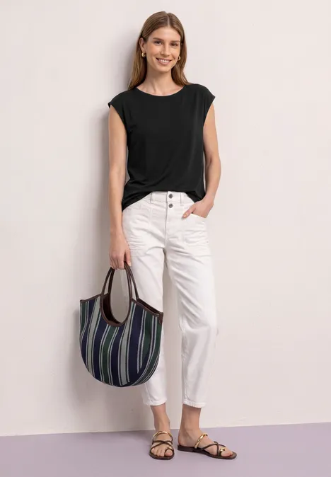 Frau in schwarzem Top, weißen Jeans und Sandalen hält eine gestreifte Tasche.