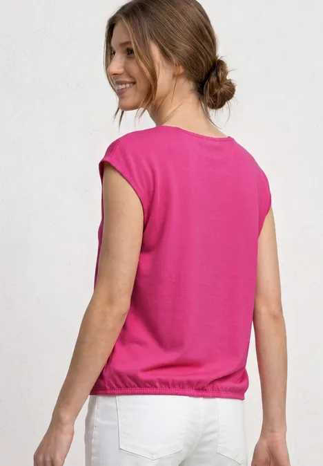 Een vrouw met opgestoken haar draagt een roze top en witte broek, ruggezicht tegen een lichte achtergrond.