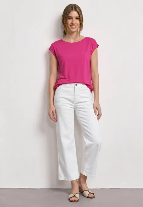 Een vrouw draagt een roze top, witte broek en sandalen, staand voor een lichte achtergrond.