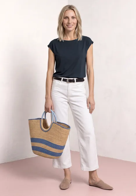 Frau in schwarzem Shirt, weißen Hosen und flachen Schuhen trägt eine gestreifte Strohtasche vor schlichtem Hintergrund.