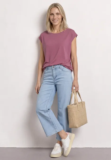 Vrouw in paarse top, lichtblauwe jeans, witte sneakers en strooien tas.