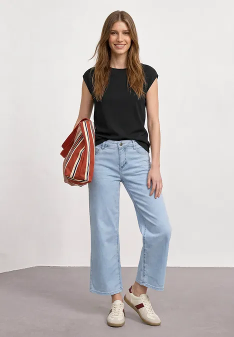 Basic shirt met boothals Black Vrouw in zwart T-shirt, lichtblauwe jeans, witte sneakers met gestreepte tas.