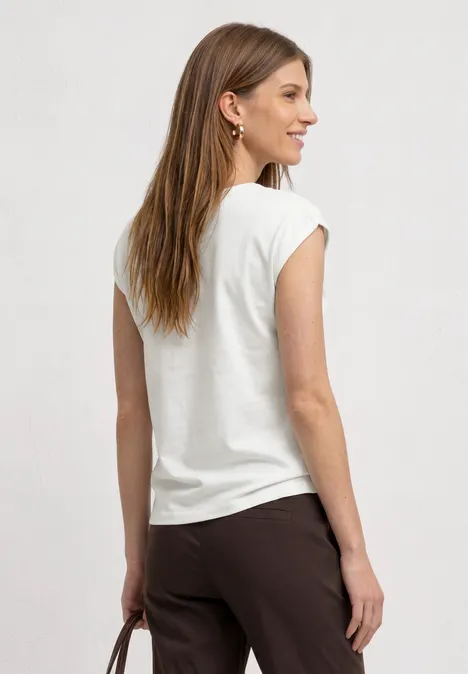 Basic shirt met boothals off white Een vrouw van opzij draagt een wit T-shirt, een bruine broek en houdt een tas vast.