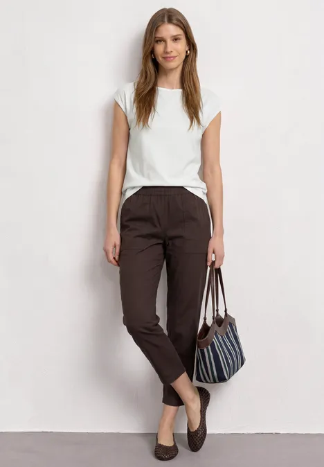 Basic shirt met boothals off white Een vrouw in casual kleding staat voor een witte muur, met witte top, bruine broek en tas.