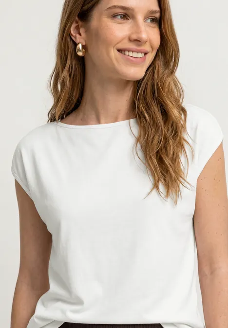 Basic shirt met boothals off white Vrouw in een witte top met gouden oorbellen, glimlacht tegen een lichte achtergrond.