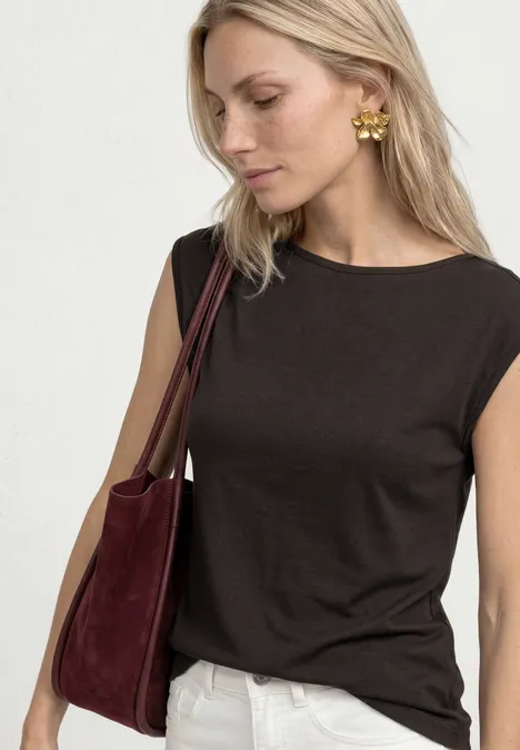 Femme blonde en haut noir sans manches, pantalon blanc, boucles d’oreilles dorées et sac bordeaux.