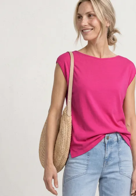Une femme porte un top sans manches rose vif, un jean clair et un sac rond en paille.