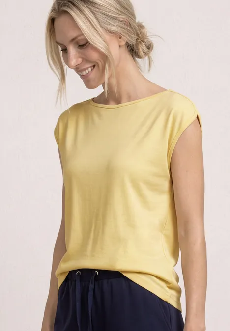 Slanke vrouw draagt een gele mouwloze top en een donkerblauwe broek tegen een neutrale achtergrond.