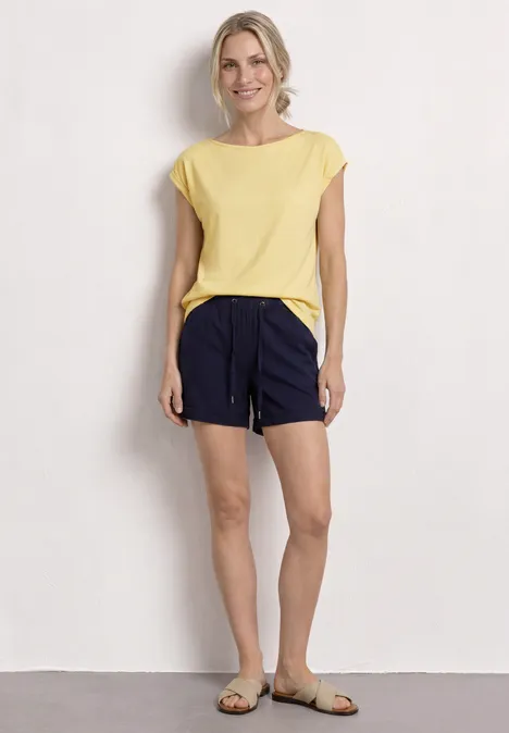 Een vrouw draagt een lichtgeel t-shirt, donkerblauwe short met trekkoord en beige sandalen voor een witte muur.