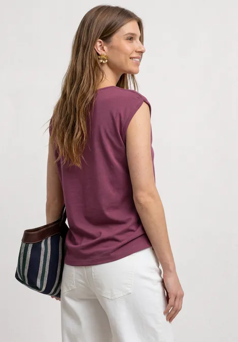 Une femme vue de dos portant un top violet sans manches, un pantalon blanc et un sac rayé.