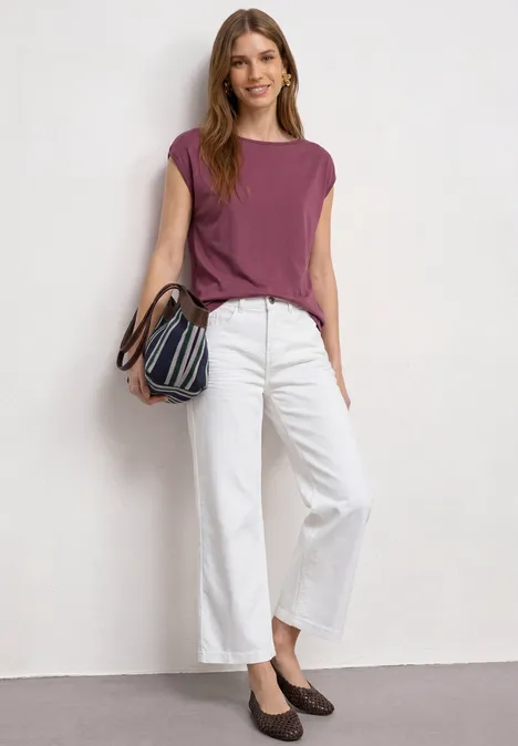 Personne en top violet, pantalon blanc large, chaussures foncées et sac rayé devant un mur blanc.